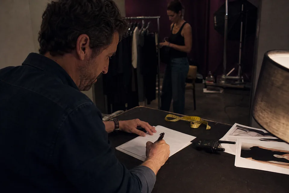 A booker's hands sign a printed model booking agreement on a dark set table beside a tape measure and a stack of comp cards, under one warm lamp