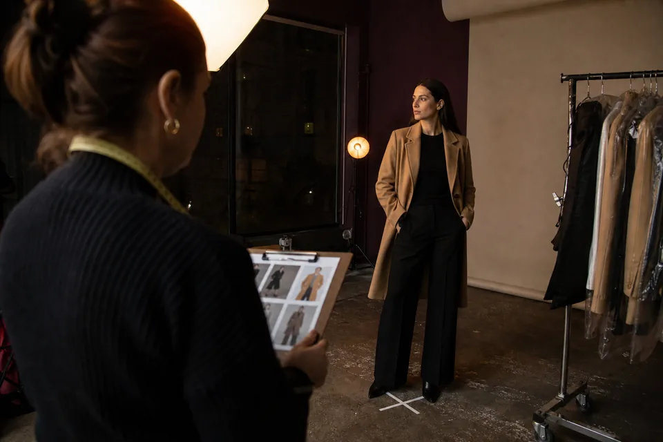 Casting day in a dim studio: a model in a tailored camel coat stands on a taped mark while a producer with a clipboard checks measurements under one softbox