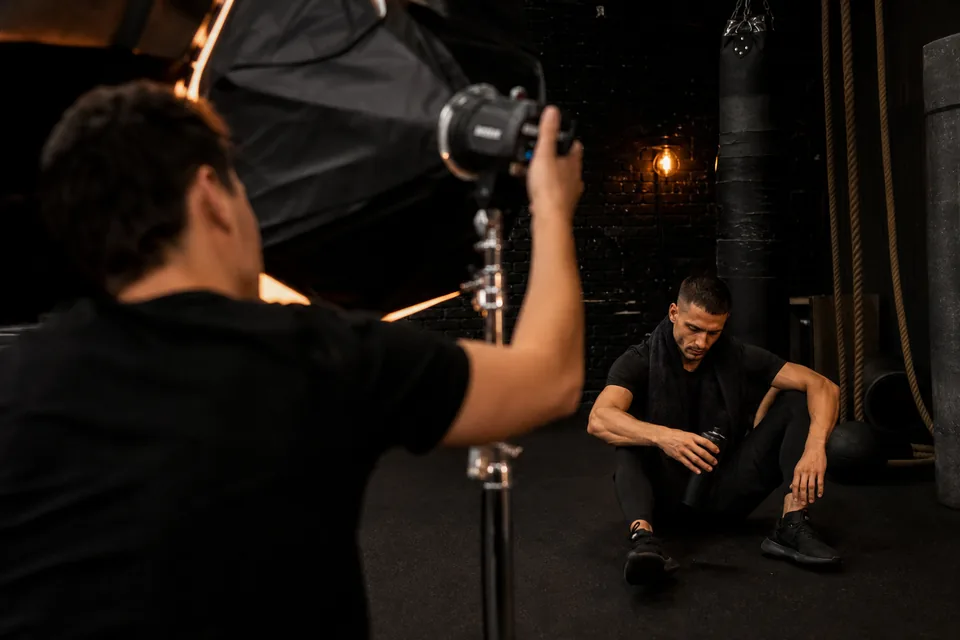 Fitness shoot: a model in matte black activewear rests between takes on a gym floor while an assistant adjusts a softbox