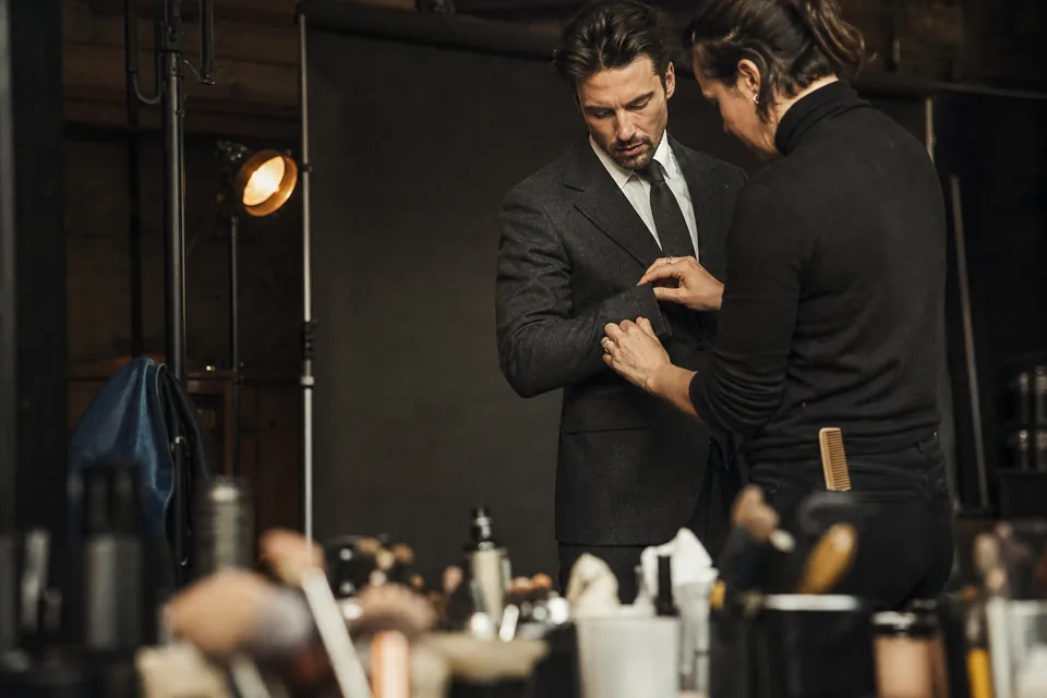 Menswear shoot: a male model in a charcoal suit is adjusted at the cuff by a groomer in front of a dark paper backdrop