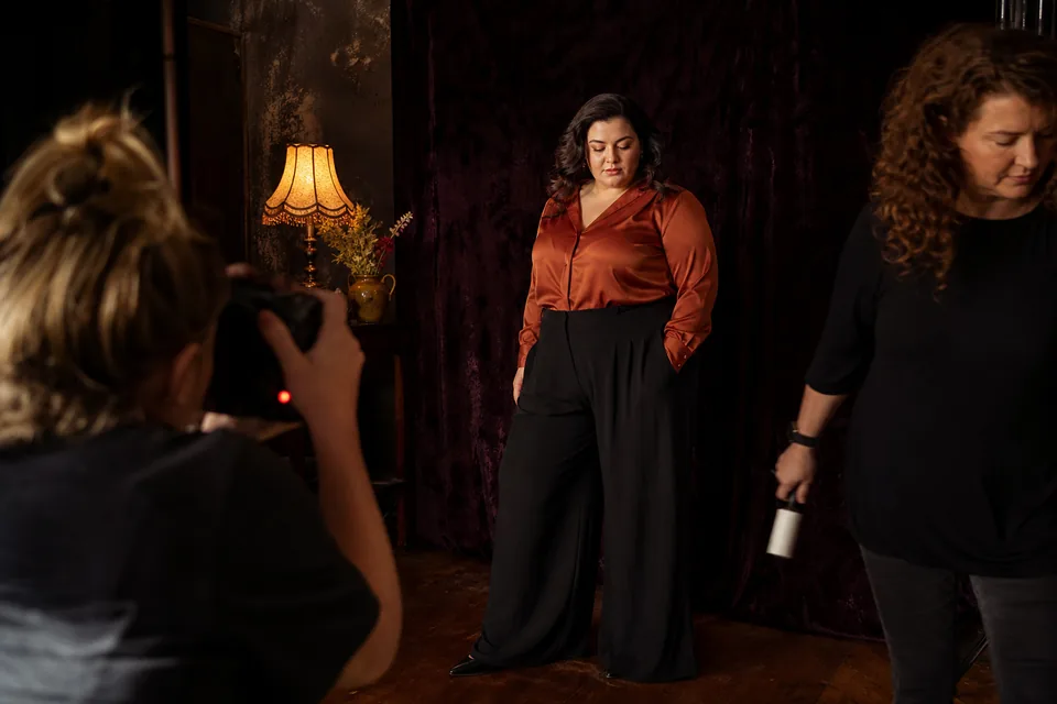 Plus size fashion shoot: a curve model in a rust satin blouse and wide trousers is photographed against a dark velvet backdrop while a stylist steps out of frame